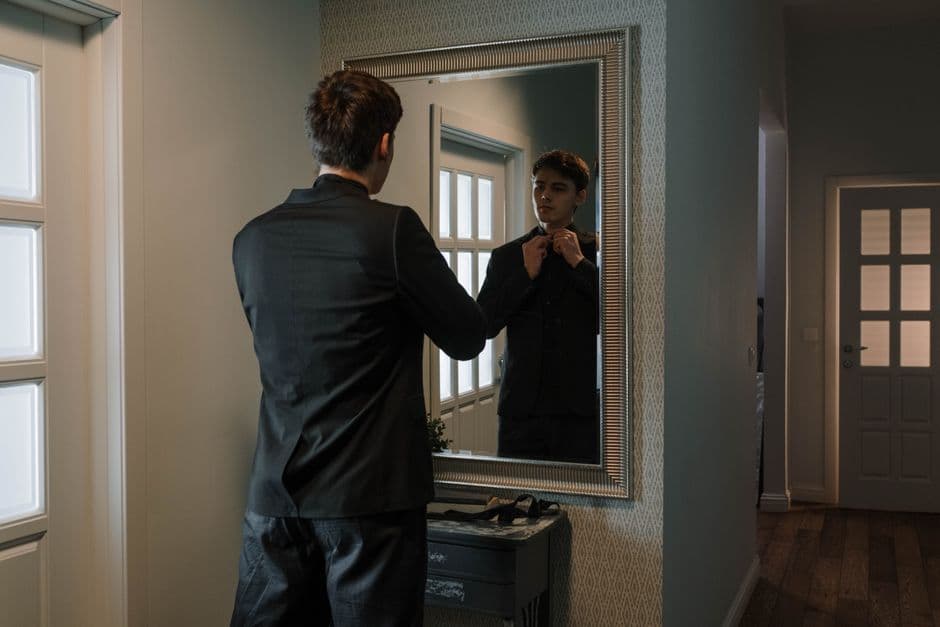A young man adjusts his suit while standing in front of a mirror in a stylish home setting.