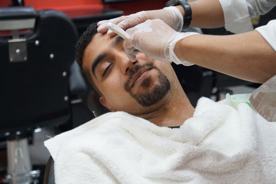 A barber carefully shaves a relaxed client using a straight razor at a salon.